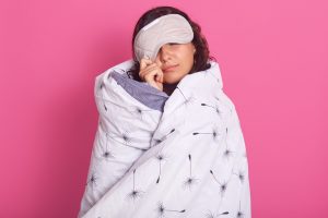 Close up portrait of brunette woman peeping from sleeping mask, wearing white blanket. For unhealthy during lockdown article