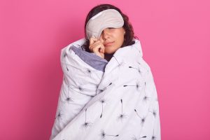 Close up portrait of brunette woman peeping from sleeping mask, wearing white blanket.  For unhealthy during lockdown article