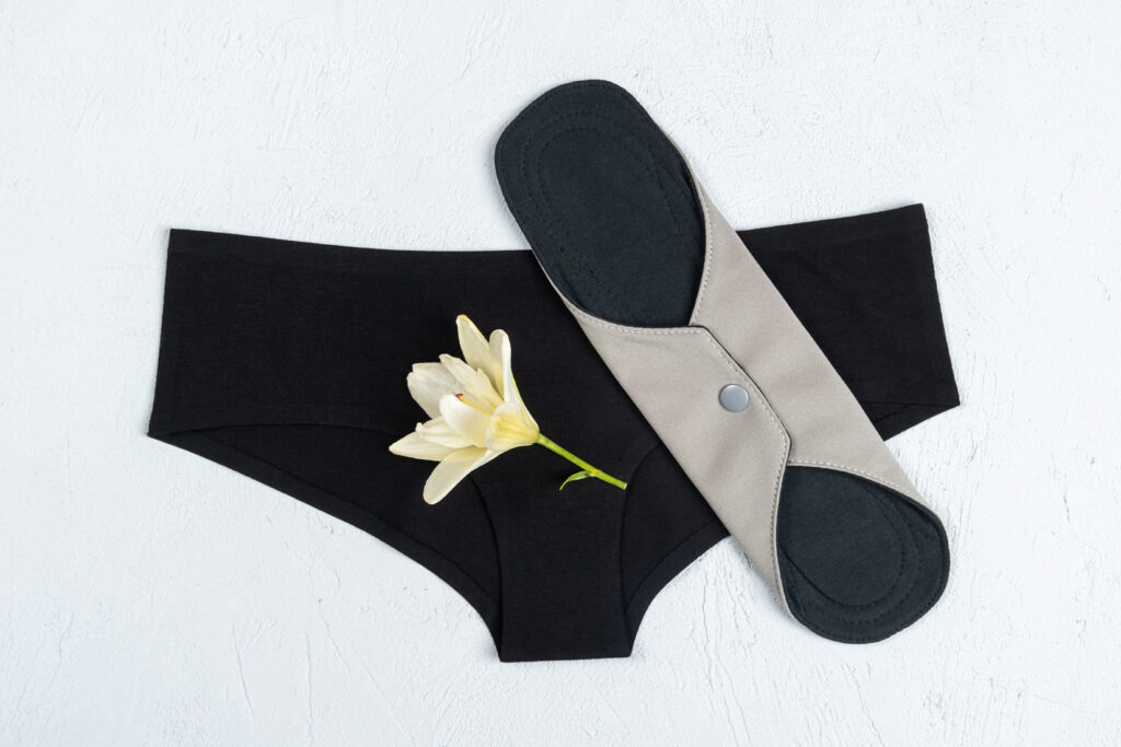 Eco sanitary reusable pad with white delicate flower and black underpants on grey background. Health care and zero-waste, no plastic concept. Top view Flat lay.
