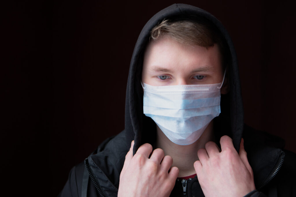 Sad, blue-eyed young man has protected himself from the coronavirus with a medical mask, and is looking down as he adjusts the hood of his jacket for Grab the Meic Fed up of Covid article.