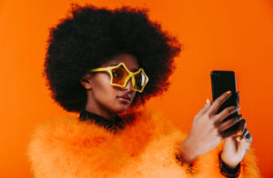 Woman with afro, star sunglasses and orange furry jacket taking selfie on phone with orange background.