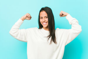 Young pretty hispanic woman showing strength gesture with arms, symbol of feminine power