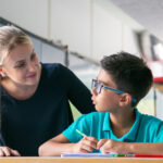 Cheerful school teacher giving help and support to schoolboy in class.