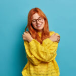 Redhead woman embraces herself expresses self love and care tilts head smiles gently closes eyes hugs own body wears warm yellow sweater expresses calmness isolated on blue wall for How To Cope With Distressing Things in the News blog