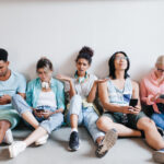 Exasperated girl with curly black hair sat on floor with group of friends on their smartphones with backs pressed against wall for What To Expect On The Day Of Exams blog