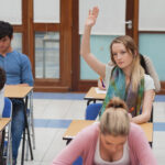 Girl raising hand during exam in exam hall for What To Expect On The Day Of Exams blog