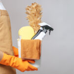 Side view, Close up hand of Young man in apron and rubber gloves holding a basket of cleaning equipment - feather duster, spray bottle, sponge and cloth.