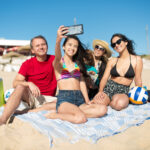 Smiling family taking a selfie at the beach sat on a towel on the sand for Things to Do After Exams blog