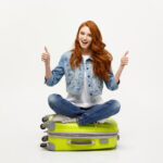 Young excited girl with long red hair sitting on a suitcase with thumbs up for Things to Do After Exams blog