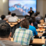 Speaker on the stage in front of the room with rear view of Audience 