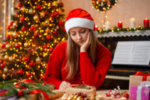 Sad young girl in red sweater and santa hat sits at the table and watches something on smartphone. Festive decoration of the room, but bad mood of person