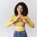 curly-haired girl showing heart sign with hands on chest smiling happily
Blog caredigrwydd