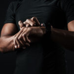 Close-up photo of african man in black t-shirt check out time at black wristwatch, over black background