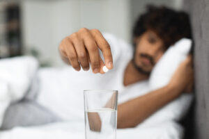 young man lying in bed, putting pills into glass of water