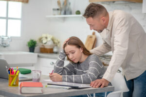 Short-haired man standing over a young girl with concerned hand on her back. Girl is trying to write with a pained expression on her face. For dyslexia blog.