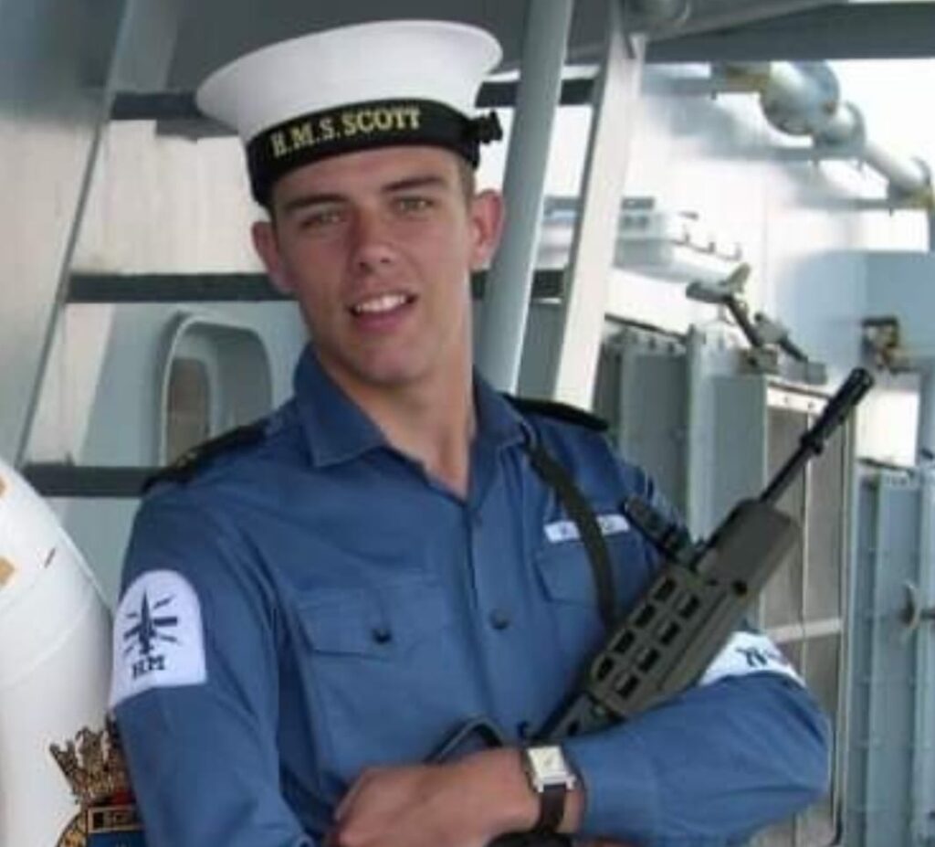 Young man smiling at camera in Navy uniform carrying a machine gun