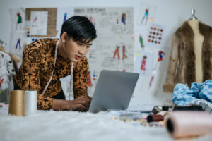 Portrait of Professional Asian young male tailor with measuring tape on neck working on laptop with drawings background in studio. Designer and LGBTQ Concept.