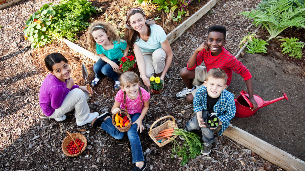 Group of children and teenagers in community garden, smiling at camera, surrounded by freshly picked produce and flowers