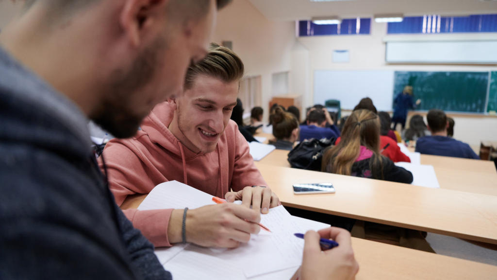 Two male students collaborate on notes in a lecture hall while others listen to the instructor at the front of the room.
University life