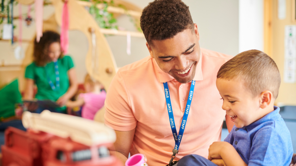 Young adult wearing a staff lanyard in a childcare setting. He is talking to a young toodler. 