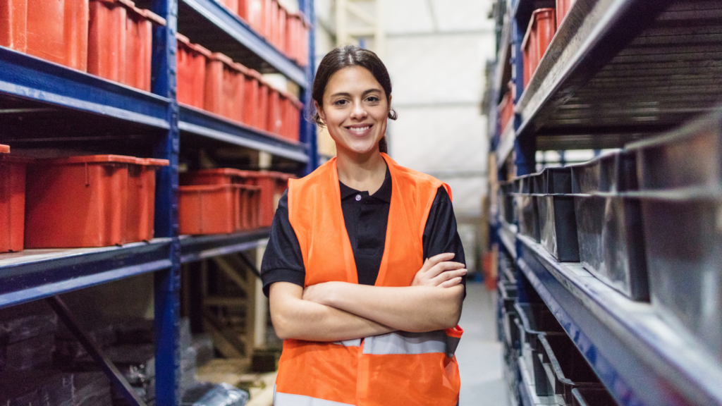 Young woman wearing an orange high visibility jacket, smiling at the camera in a warehouse.