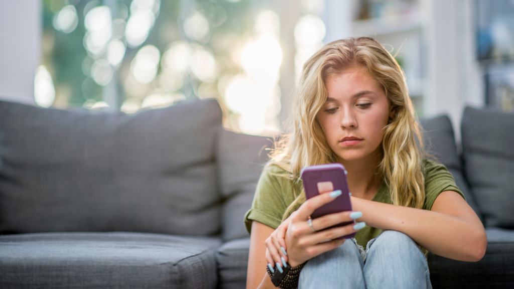 Girl sat on the floor leaning against the sofa. She is looking down at her mobile phone.