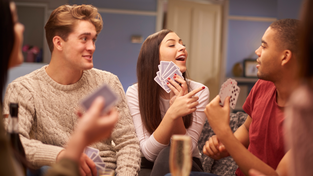 Group of 5 young adults sat around a table filled with alcohol drinks, playing a card game. One girl points at a boy, whilst another boy watches and laughs. 