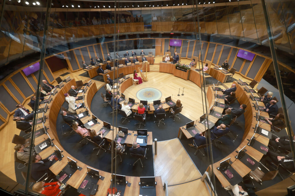 Llun o siambr crwn y Senedd o'r top i lawr. Aelodau'r Senedd yn eistedd wrth y desgiau. 