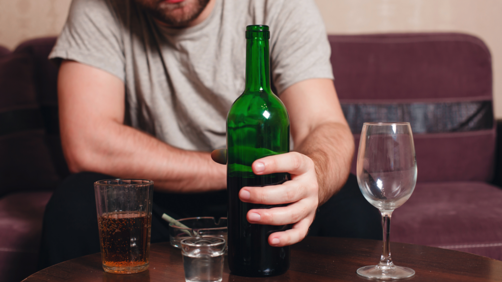 Man sat alone on sofa drinking alcohol. He has a bottle of wine in his hand, an empty wine glass, and a shot glass on the table.
