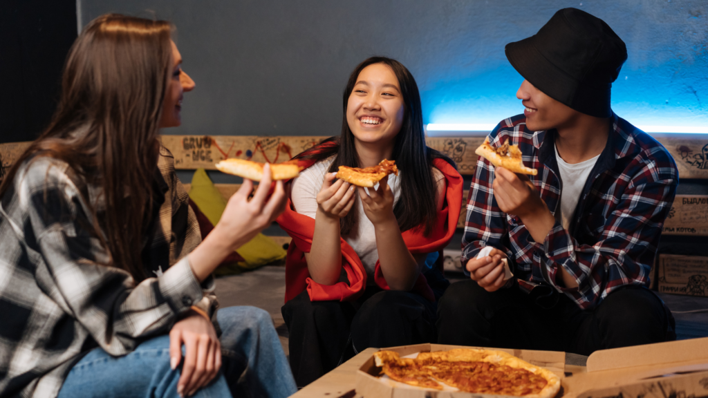Three young people smiling whilst eating slices of takeaway pizza around a table