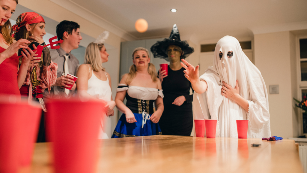 7 young people stood around a large wooden kitchen table, playing a game of beer pong and holding red solo cups with alcohol in them. They are dressed in halloween costumes at a party, with one young person dressed as a ghost, and others being a witch, angel, devil and pirates. 