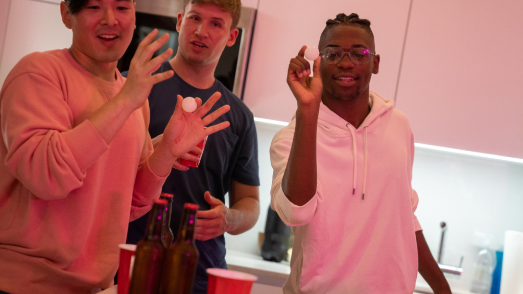 Three young adult men playing a game of beer pong on the table. There are bottles of beer and other alcohol around the room. 