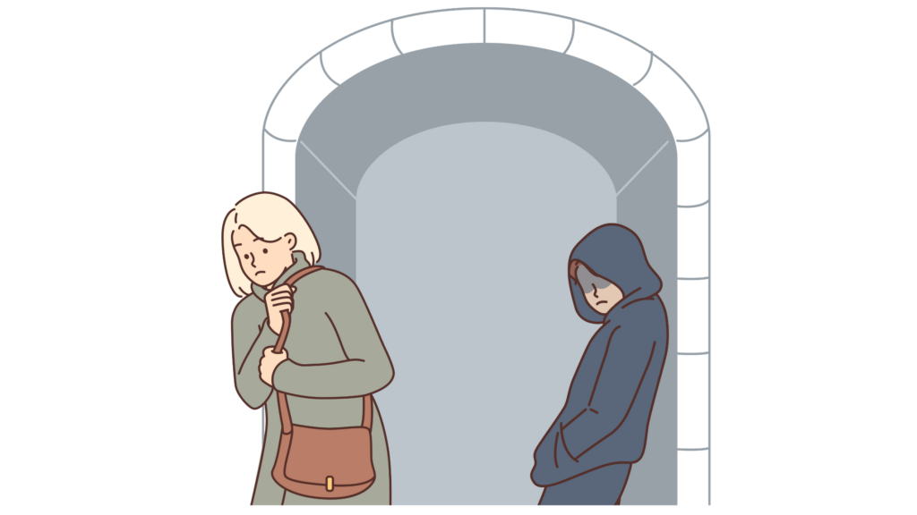 Woman walking in the street wearing a coat and a cross-body bag. There is a man in the dark in the background has his hood up and a mask on. He is following the lady.