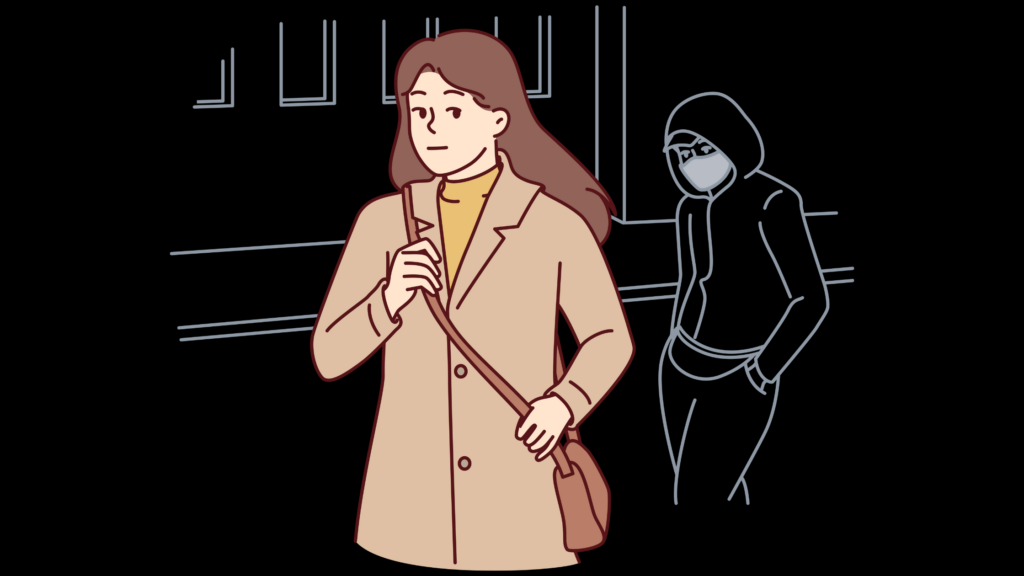 Woman walking in the street wearing a coat and a cross-body bag. There is a man in the dark in the background has his hood up and a mask on. He is following the lady.