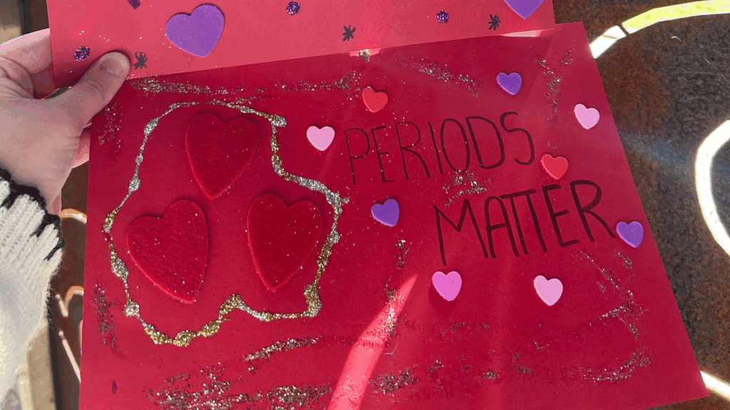 Homemade banner with with red paper, glitter and heart stickers. The poster reads 'periods matter'.