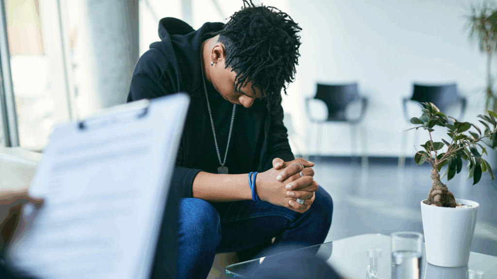 Teen boy sat in a counselling therapy  room holding his hands together with his head down